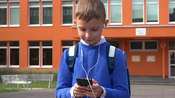 A Young Boy Listens To Music on a Smartphone with Earphones - an Elementary School in the Background alt
