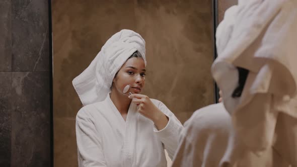 Young Darkskinned Brunette in Front of a Mirror in a Robe Massages Her Face alt