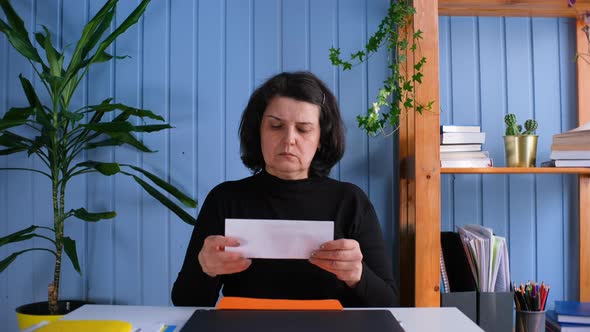 Middle Aged Woman Sit at Workplace Desk Holding Papers Reading Bad News