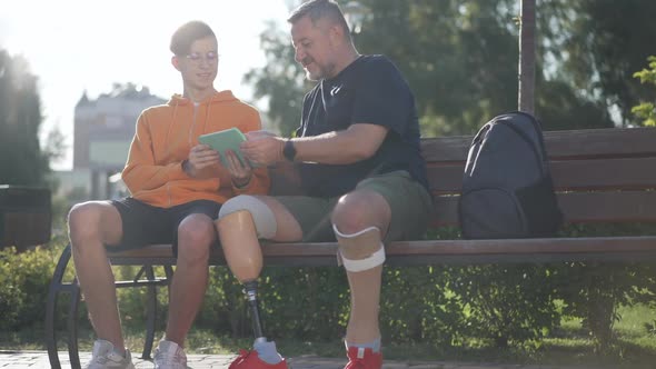 Smiling Mature Male Amputee and Teenage Boy Talking in Slow Motion Sitting in Sunbeam on Bench in alt