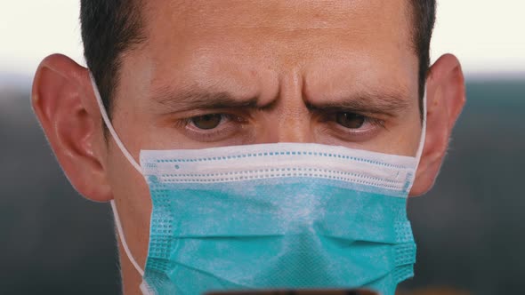 Man in Protective Face Mask Looks at Smartphone Holding It in Hands on Street alt