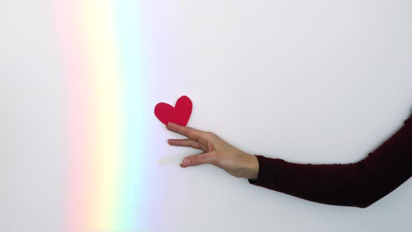 Female hand holding red paper heart on the rainbow background.