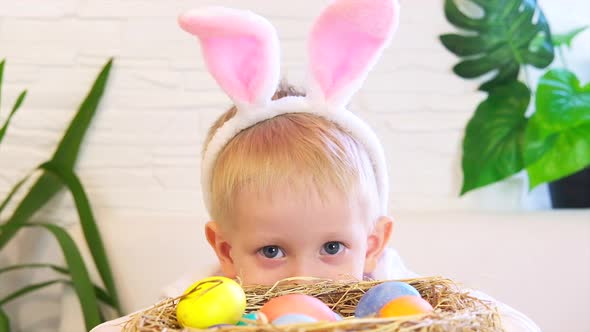 A child boy with rabbit ears peeks out of a basket of easter eggs and looks at the camera