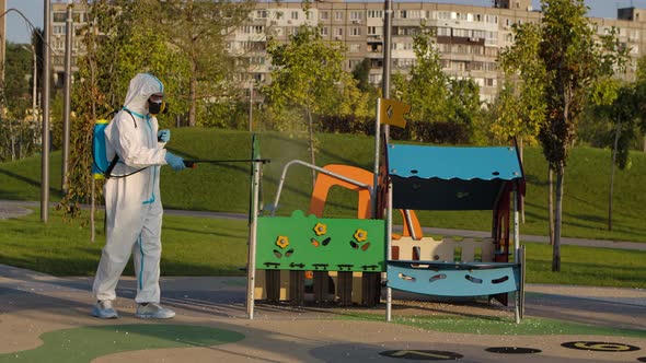 A Man in a Protective Suit and a Respiratory Gas Mask Disinfects the Slides on the Playground alt