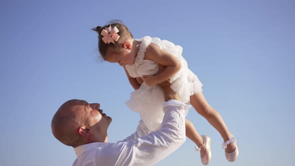 Side View of Confident Cheerful Father Raising Cute Little Girl in Dress at Background of Clear Blue alt