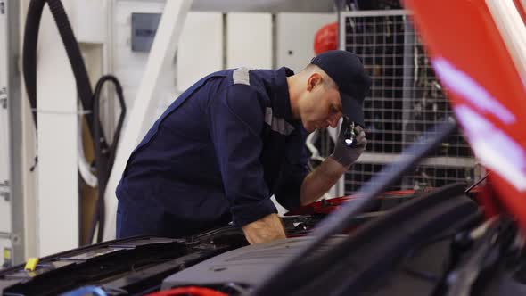 Car Service Worker Examinating Engine Under Hood with Flashlighter in Hand Side View alt