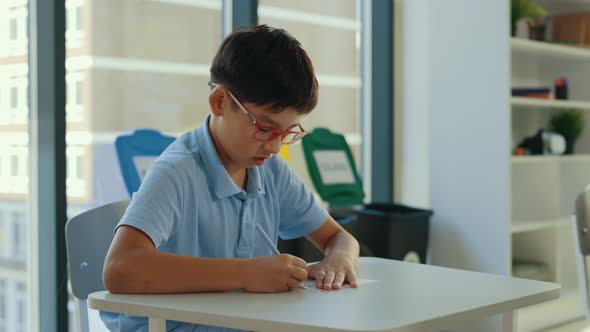 Asian Pupil Wearing Glasses Sits at a Desk at School and Fills Out Tests alt