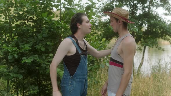 A Guy in a Farm Jumpsuit Puts a Straw Hat on a Guy in a Tshirt