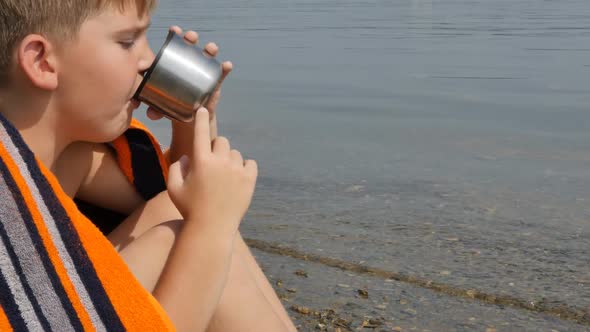 a Teenage Caucasian Boy Sits on the Shore of the Lake Wrapped in a Warm Towel and Drinks Tea From a alt
