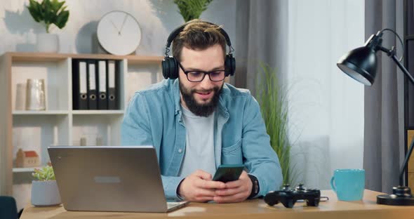 Bearded Man in Headphones Enjoying Music from Phone Playlist Sitting at His Workplace at Home alt