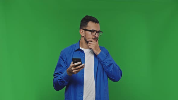 Attractive Guy Texting on His Phone Scratching His Beard Thinking Looking at Camera and on Green alt