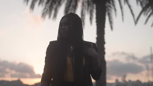 Gorgeous black woman touching long braids near palm in twilight alt