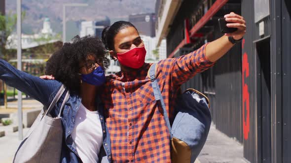 Diverse couple wearing face masks embracing and taking a selfie alt