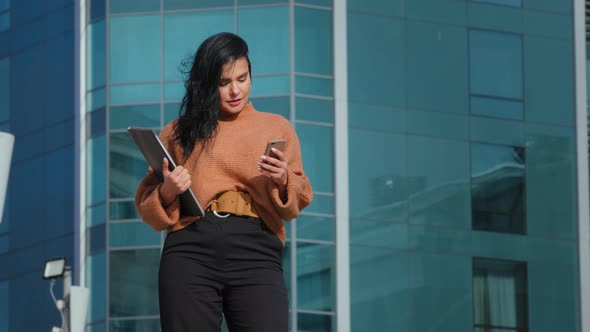 Young Hispanic Businesswoman Stands Backdrop Office Building Looks at Telephone Screen Sending Email alt