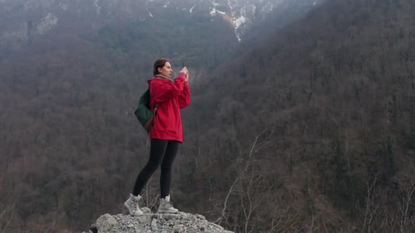 A Girl in a Red Raincoat Walks and Takes Photos of the Mountains