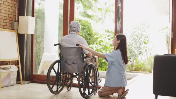 Old senior Asian man sitting in wheel chair in nusing home looking out of window alt