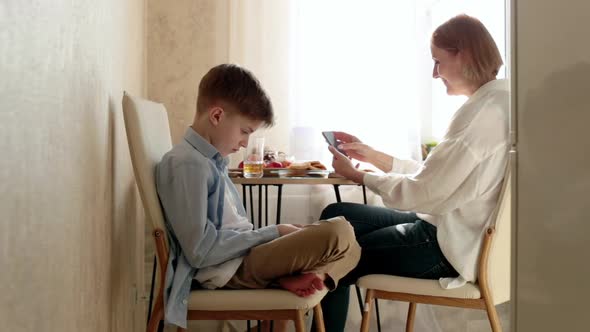 Woman and Child are Sitting at Table in Kitchen and Looking at Their Phones