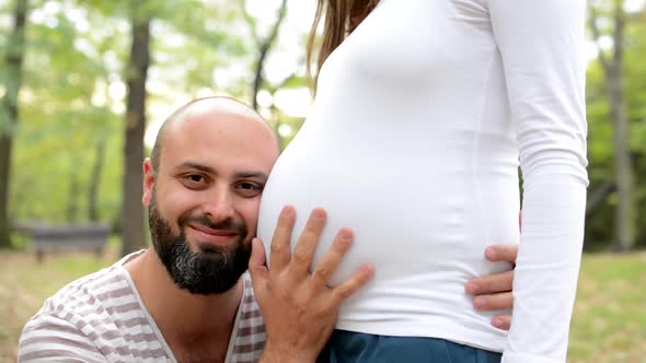 Man Strokes Abdomen of a Pregnant Woman and Couple Smile To Camera - Man Listens Abdomen in Park alt