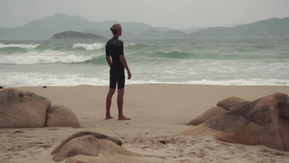 Surfer Looks at Raging Waves Standing on Sandy Beach