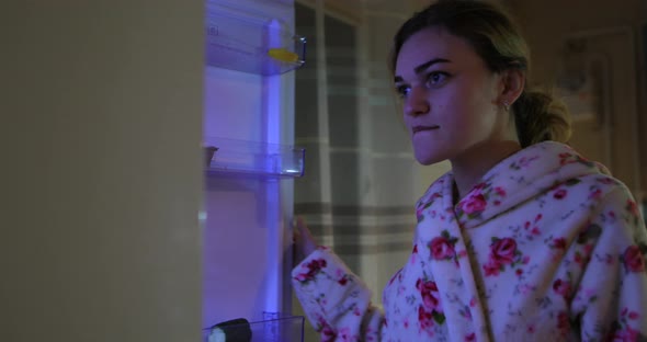 Young Woman Opens the Refrigerator at Night and Takes Food