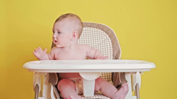 Mother feeding toddler baby from a spoon on a high chair for children, studio yellow background alt