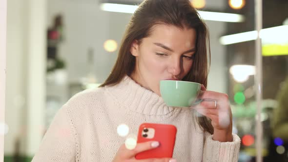 Caucasian Cheerful Woman Scrolling on Smartphone and Drinking Green Tea in Cozy Cafe