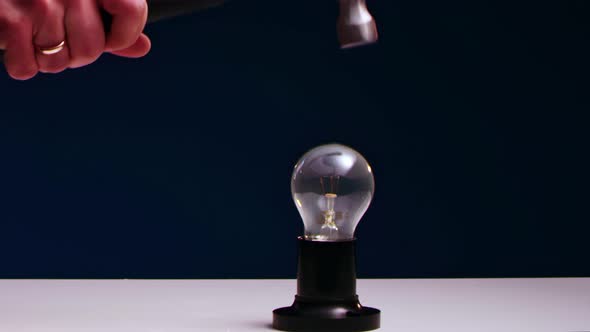 AMan's Hand Smashes Incandescent Light Bulb Lying on Table with Iron Hammer