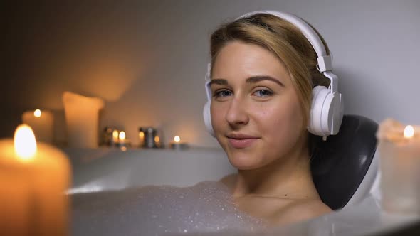 Smiling Girl in Headset Lying in Bath With Foam and Candles, Looking at Camera alt