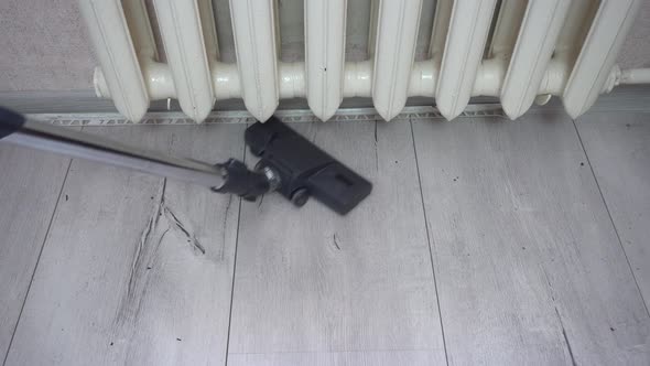 The Vacuum Cleaner Brush Cleans the Gray Wood Floor Near the Heater Radiator alt