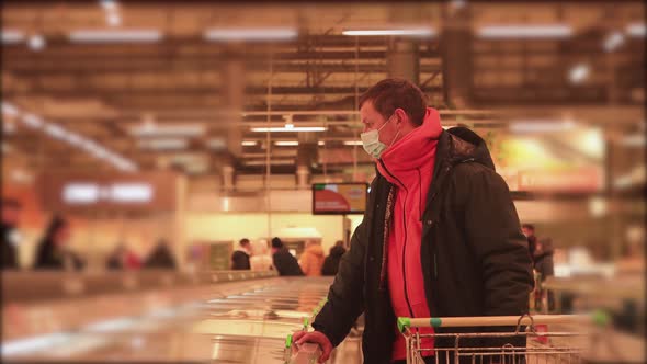 A Man with a Trolley in a Supermarket Looks at Prices with a Mask on His Face alt