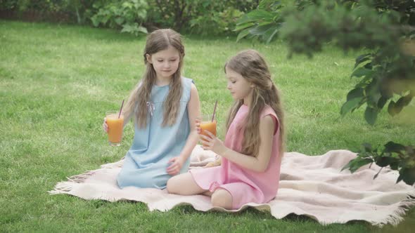Wide Shot of Curly-haired Twins in Blue and Pink Dresses Drinking Orange Juice Outdoors. Portrait of alt