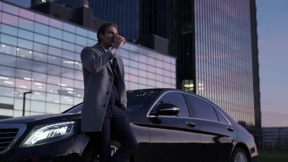 Evening Time Portrait of a Cheerful Businessman Standing Near a Business Car and Drinking Coffee Top alt