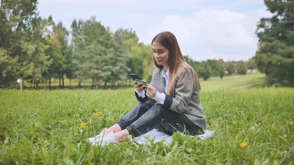A Young Girl Makes a Purchase with Her Phone and Bank Card in the Park Sitting on the Grass alt