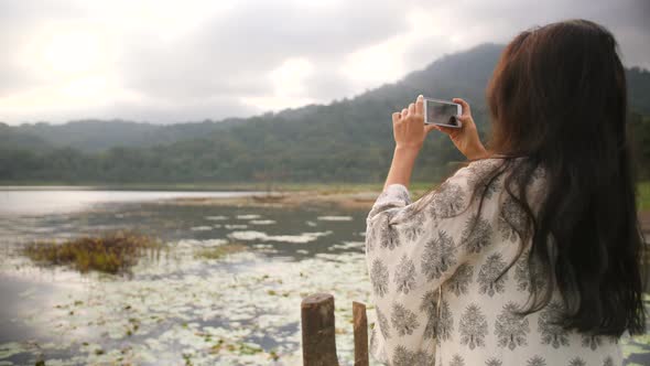 Young Mixed Race Traveler Girl Taking Photo of Beautiful Morning Lake and Mountains View Using alt