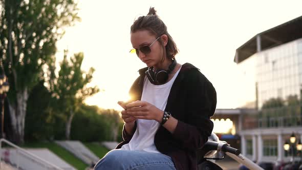 A Fashionable Modern Girl Sits on a Moped and Texting on Her Mobile Phone alt