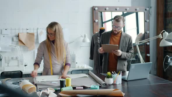 Man and Woman Sewing Studio Workers Talking Choosing Material and Using Laptop alt