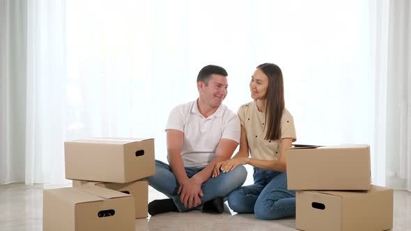 Young Happy Couple Sitting on the Floor of Their New Apartment Among Cardboard Boxes alt