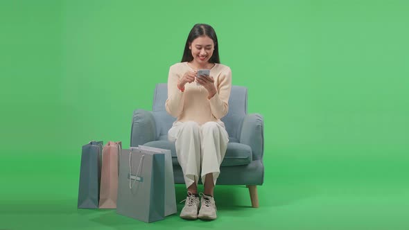 Front View Of Asian Woman Sitting On Sofa With Shopping Bags Enjoy Using Mobile Phone On Green Scree alt