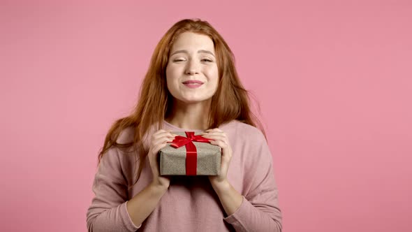 Cute Young Woman Holding Gift Box with Bow, She Wonders What Is Inside. Pink Wall Background. Girl alt