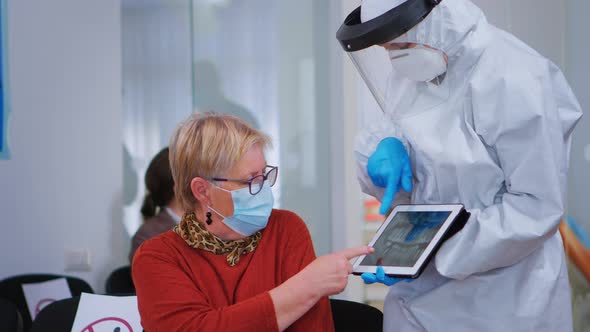 Woman Sitting on Chair Listening Doctor with Coverall Looking on Tablet alt
