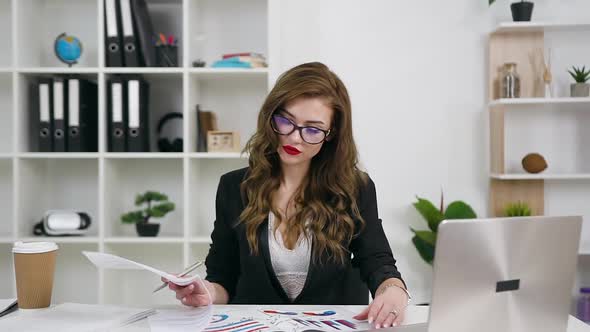 Businesswoman in Glasses in Stylish Wear and with Bright Make-up which Reviewing Reports with Graphs alt