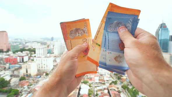 A Young Man Recounts in His Hands the Money of Malaysia Against the Background of the City Center alt