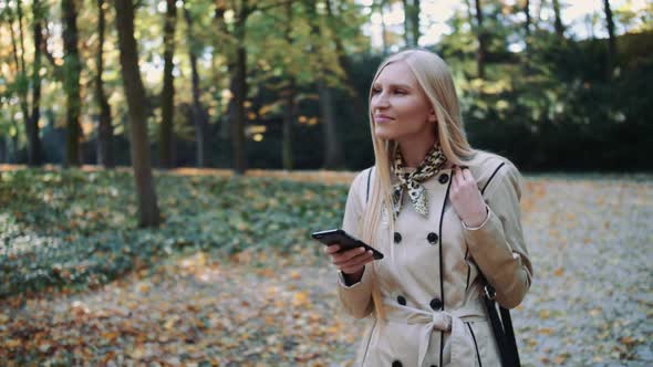 Young Girl Walking in Autumn Park and Texting a Message on a Mobile Phone alt