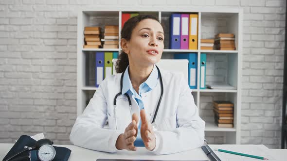 Doctor Female in White Uniform with Stethoscope is Consulting Someone Sitting at Table in Cabinet of alt
