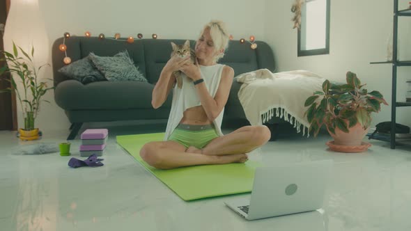 Mature Woman Sits with Cat on Yoga Mat in Front of Laptop in Preparation for Online Yoga Class alt