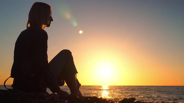 woman sitting on pebble beach by sea and throwing stones into water. alt