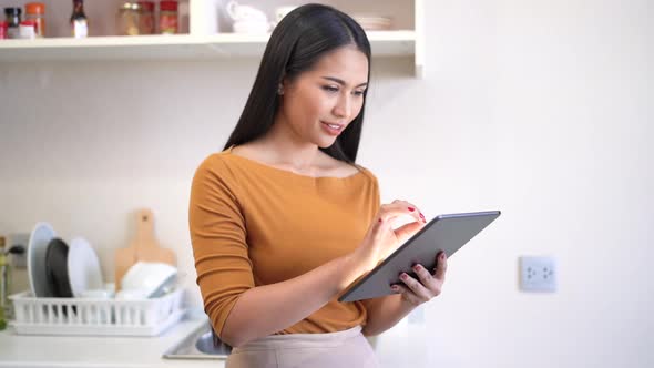 Young woman relaxing standing in kitchen and using digital tablet.