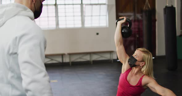 Fit caucasian woman wearing face mask working out with kettle bells with male trainer at the gym alt