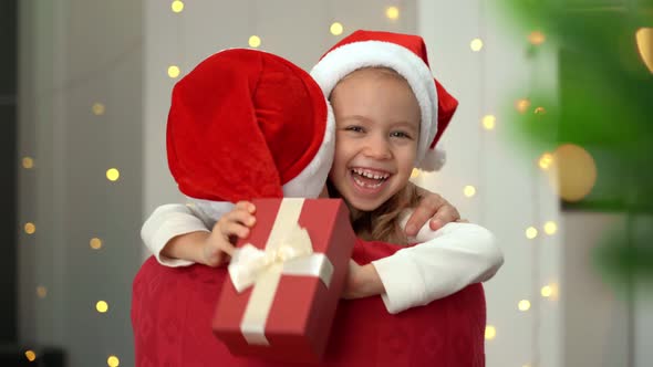 Little Girl Runs to Father with Gift Box Hugs and Laughs on Background of Christmas Tree