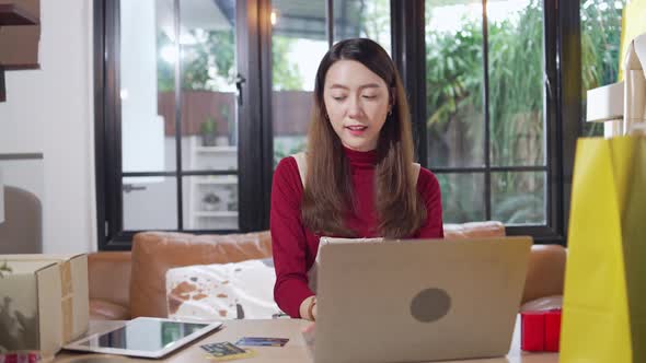 Asian female shop owner checking customer order on digital tablet and managing stock on laptop alt
